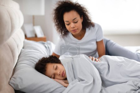 Un moment tendre alors que la mère afro-américaine observe amoureusement son enfant endormi sous une couverture confortable, avec la lumière du soleil filtrant dans la chambre sereine, suggérant un début paisible de la journée.
