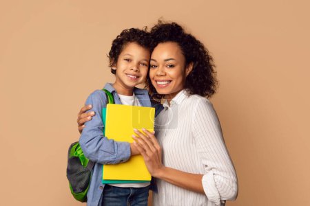 Foto de Una alegre madre afroamericana abraza amorosamente a su hijo pequeño, que está preparado para un día en la escuela con su mochila y un cuaderno amarillo brillante - Imagen libre de derechos