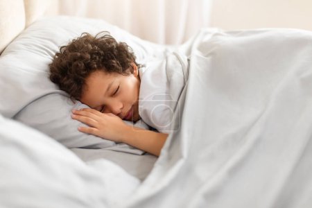 Ein junger afroamerikanischer Junge schläft friedlich in einem Bett, das mit knackig weißen Laken bedeckt ist. Sein Kopf ruht auf einem flauschigen Kissen, während er Ruhe und Trost in seiner Umgebung findet..
