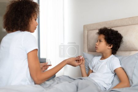 Téléchargez les photos : Une mère afro-américaine inquiète s'assoit sur le bord d'un lit, tenant doucement la main de son jeune enfant pendant qu'elle vérifie s'il y a de la fièvre. L'enfant a l'air malade. - en image libre de droit