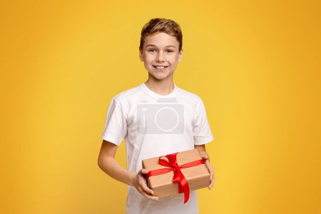 Foto de Un niño de pie sosteniendo una caja de regalo envuelta con una cinta roja, presentándola al espectador. - Imagen libre de derechos