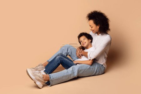 Foto de La joven madre afroamericana se sienta en el suelo, acunando a su hijo en sus brazos con un cálido abrazo. El fondo es un beige liso y suave - Imagen libre de derechos