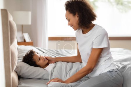La madre afroamericana toca suavemente la cara de su hijo mientras comparten un momento amoroso en una mañana serena. Confort y afecto resuenan en este apacible comienzo del día.