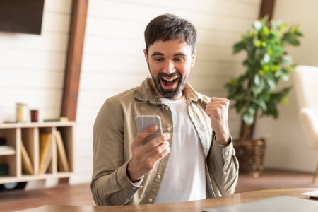 A young man is visibly overjoyed while looking at his smartphone, pumping his fist in a gesture of victory or celebration. The indoor setting suggests a personal or professional triumph