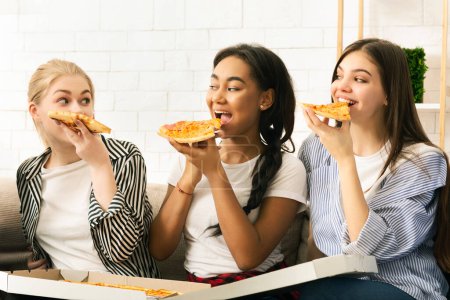 Téléchargez les photos : Trois jeunes filles sont assises sur un canapé, dégustant des tranches de pizza. Les filles sont vêtues de façon décontractée et semblent passer un moment détendu et agréable ensemble. - en image libre de droit