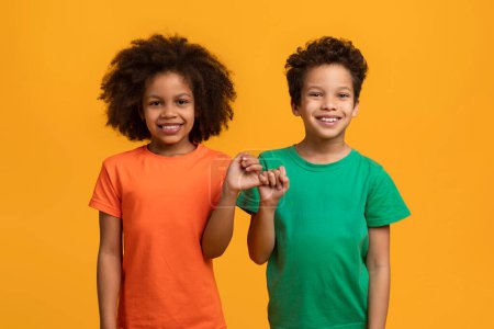 Photo for African American young boy and girl are standing side by side, holding hands with bright smiles on their faces, both wearing colorful t-shirts, standing against a vivid yellow backdrop - Royalty Free Image