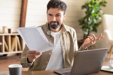 Un hombre con barba muestra una mirada de confusión o preocupación mientras examina los papeles en sus manos. Se sienta en un ambiente informal de oficina en casa con una computadora portátil abierta frente a él