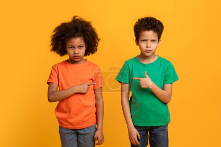 African American boy and girl, presumably siblings, stand side by side with puzzled looks on their faces, each pointing at the other against a solid yellow backdrop