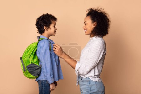 Téléchargez les photos : La femme afro-américaine aide un jeune garçon à mettre son sac à dos, à se préparer pour un voyage ou une école. Les mains de la mère ajustent doucement les sangles sur le sac à dos des garçons pour assurer un ajustement approprié. - en image libre de droit