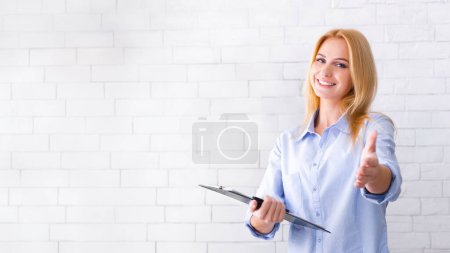 A smiling professional businesswoman stands against a white brick wall holding a clipboard in one hand while extending her other hand for a handshake, copy space