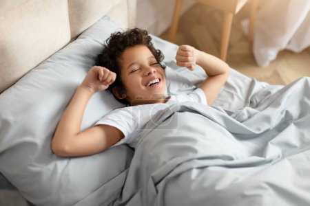 Photo for African American young boy is lying comfortably in his bed, with a soft smile on his face. He looks content and relaxed, enjoying a moment of peace before sleep, stretching - Royalty Free Image