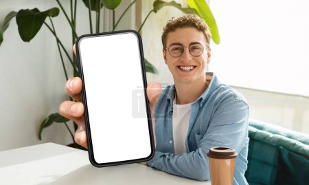 Téléchargez les photos : Un jeune homme joyeux avec des cheveux bouclés et des lunettes est assis à une table au café, montrant un smartphone avec un écran vide vers la caméra - en image libre de droit
