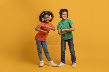 African American young boy and girl stand side by side, each striking a playful pose with their arms while dancing, using wireless headphones, listening to music