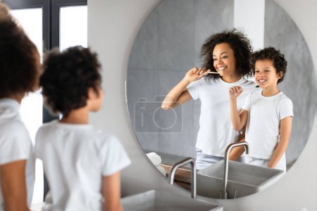 Foto de Una joven y alegre madre afroamericana le muestra a su hijo cómo cepillarse los dientes correctamente en un baño iluminado. Tanto la madre como el niño están de pie frente al fregadero, usando cepillos de dientes. - Imagen libre de derechos