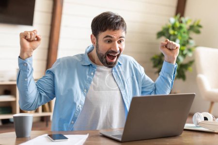 Foto de Un hombre alegre en una camisa azul casual está levantando sus puños en triunfo delante de su computadora portátil, su cara expresando euforia, un ambiente de oficina en casa lleno de luz natural. - Imagen libre de derechos