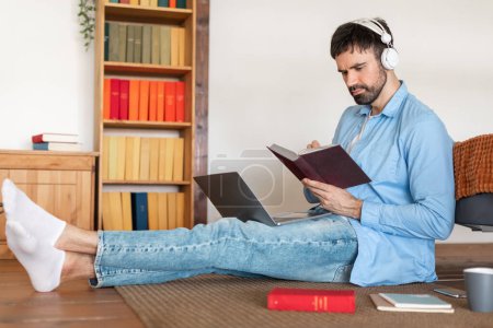 Un hombre enfocado se sienta en una alfombra con un portátil en su regazo y un cuaderno en la mano, con auriculares. Una taza de café, un teléfono inteligente y libros adicionales lo rodean