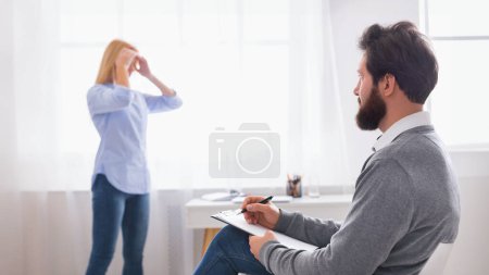 Man therapist is seated, holding a notepad and pen, facing woman client who appears to be in distress, with her hand on her head, in therapy office, emotional support.