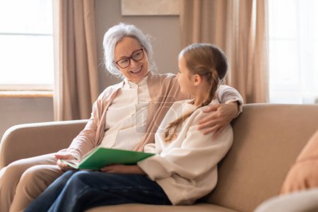 Photo for An elderly woman with glasses and a young girl are sharing a moment of bonding while sitting on a comfortable couch, woman is holding a book, suggesting she is reading to her attentive granddaughter - Royalty Free Image