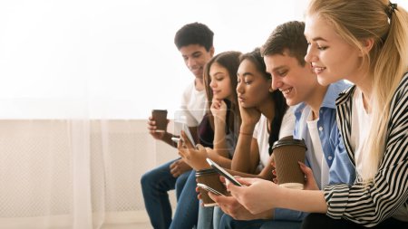A group of multiethnic teenagers is seated close to one another, each engrossed in their own smartphones. They appear to be in a relaxed social setting