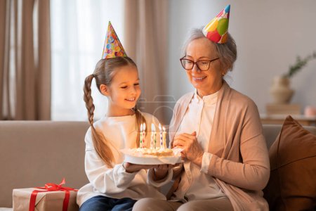 Téléchargez les photos : Une femme plus âgée et une jeune fille s'assoient sur un canapé, souriant à un gâteau d'anniversaire devant eux. La femme allume des bougies sur le gâteau tandis que la fille attend avec impatience une tranche. - en image libre de droit