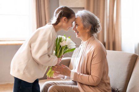 Téléchargez les photos : Une jeune femme présente tendrement un bouquet de tulipes blanches fraîches à une dame âgée, qui les reçoit avec une expression joyeuse, assise dans un salon chaleureusement éclairé - en image libre de droit