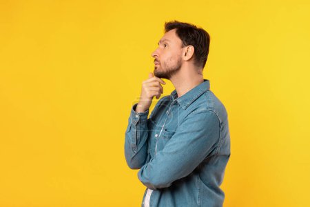 A man standing upright in front of a solid yellow background. The man is facing forward with a neutral expression, touching his chin and looking at copy space
