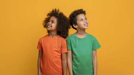 Two joyful African American children are standing side by side, one wearing an orange shirt and the other in green, both gazing upwards with happy expressions on their faces