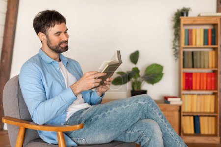 Un hombre está sentado en una silla, absorto en la lectura de un libro. La atención de los hombres se centra en las páginas del libro, su postura relajada como él da vuelta a través de las páginas.