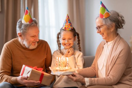 Foto de En una habitación cálida y acogedora, una joven está encantada con un pastel de cumpleaños encendido presentado por su abuela sonriente, mientras que un abuelo feliz mira y sostiene un regalo envuelto - Imagen libre de derechos