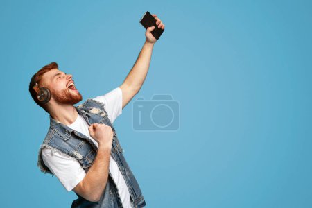 Handsome guy emotionally singing with closed eyes, listening music in headphones on blue background, copy space