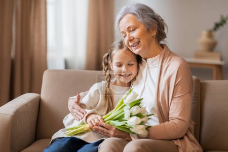 Foto de Un momento tierno es capturado mientras una abuela sonriente envuelve sus brazos alrededor de su nieta joven, que sostiene un ramo de tulipanes frescos - Imagen libre de derechos