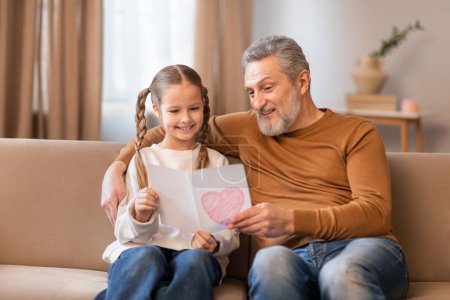 A grandfather and his young granddaughter are sitting together on a beige sofa, sharing a special moment. The girl, with braided hair, showing a drawing of a pink heart, radiating pride and happiness