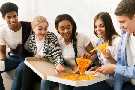 Téléchargez les photos : Un groupe d'amis assis sur un canapé, dégustant des tranches de pizza. Le groupe diversifié s'engage dans une conversation occasionnelle tout en savourant leur repas ensemble. - en image libre de droit