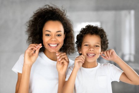 Una alegre madre afroamericana y su hijo pequeño están de pie juntos en un baño bien iluminado y contemporáneo, con camisetas blancas mientras ambos se limpian los dientes con hilo dental