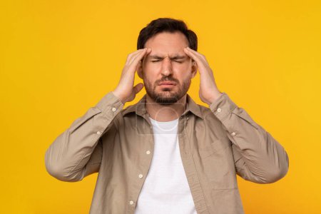 Ein Mann steht mit geschlossenen Augen und schmerzverzerrtem Gesichtsausdruck und drückt seine Finger gegen die Schläfen, was auf starke Kopfschmerzen oder Migräne hindeutet. Seine lässige Kleidung und der leuchtend gelbe Hintergrund