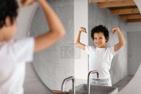 Foto de El niño afroamericano está de pie frente a un espejo del baño, orgullosamente flexionando sus músculos y exhibiendo una sonrisa alegre. El baño tiene un diseño moderno con vigas de madera visibles - Imagen libre de derechos