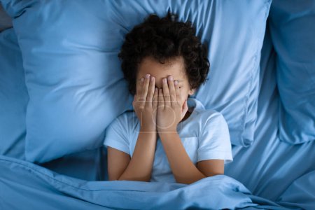 Foto de Niño afroamericano está acostado en una cama, con las manos cubriendo su cara. Parece estar profundamente en el pensamiento o experimentar emociones como la tristeza o la frustración. - Imagen libre de derechos