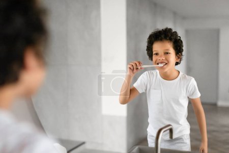 African American young boy is standing in front of a mirror, diligently brushing his teeth. He is focused on his reflection, holding a toothbrush and toothpaste