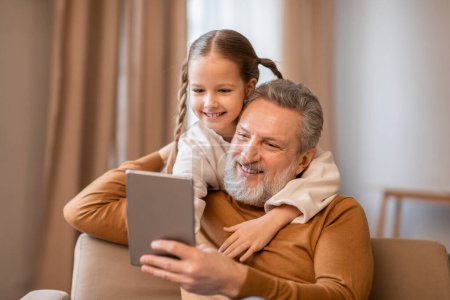Photo for An older man and a young girl are sitting side by side on a couch, both engrossed in the content on a tablet. They appear focused and engaged - Royalty Free Image