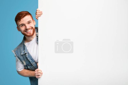 Glücklich lächelnder Typ, der aus einem weißen Blanko-Plakat blickt, das zur Werbung bereit ist, blauer Hintergrund