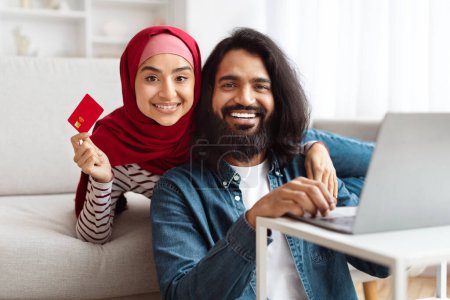 Photo for A cheerful bearded Indian man and a woman wearing a red hijab are seated together on a sofa, looking towards the camera, the man holding a laptop. The woman is showing a red credit card - Royalty Free Image