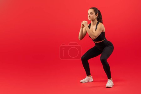A focused young woman wearing a black sports outfit with white sneakers is engaged in a squatting exercise. Her hands are clenched together in front of her as she maintains a proper form, copy space