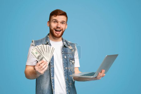 glücklicher junger Kerl, der am Laptop arbeitet und einen Haufen Geld auf blauem Hintergrund zeigt, Kopierraum