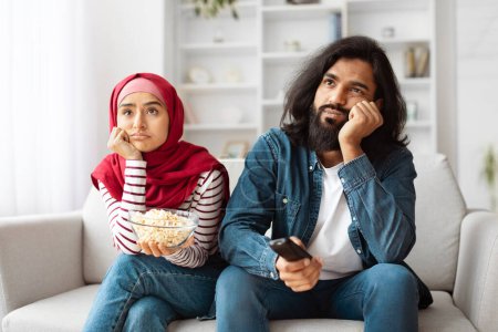 Unzufriedenes indisches junges Paar lümmelt auf einem gemütlichen Sofa in einem gut beleuchteten Wohnzimmer, scheinbar unbeeindruckt oder gelangweilt, während es eine Schüssel Popcorn in der Hand hält.