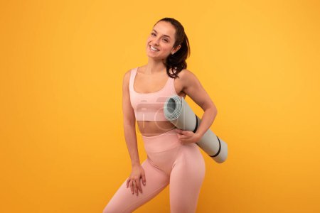 Eine Frau steht und hält eine Yogamatte in der Hand, trägt ein pinkfarbenes Sport-BH-Top und Leggings. Sie ist bereit für Yoga oder Fitnesstraining.