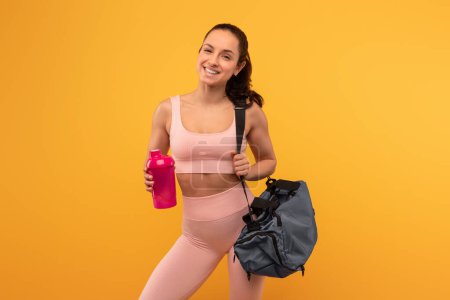 Une jeune femme joyeuse vêtue d'un soutien-gorge de sport rose et de leggings se tient en confiance sur une toile de fond jaune vif, tenant une bouteille d'eau dans une main et lançant un sac de sport sur son épaule