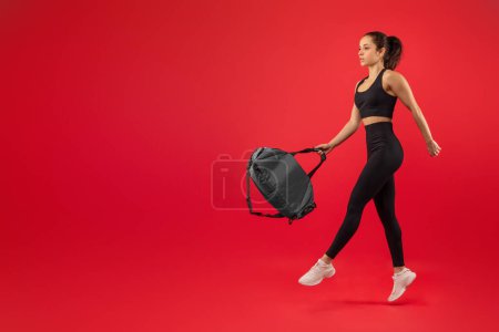 A young woman dressed in black athletic wear confidently takes a stride, holding a sports bag in one hand against a vibrant red background, copy space