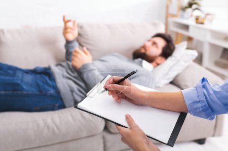 Photo for A man is lying on a couch, gesturing with his hand as he speaks, while engaged in a counseling session. A therapist, partially visible in the foreground, is attentively taking notes on a clipboard. - Royalty Free Image