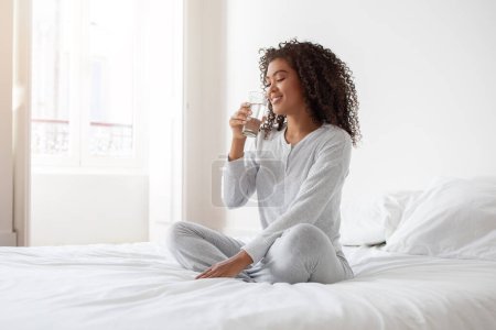 La mujer hispana está sentada en una cama, sosteniendo una copa de vino tinto. Ella está vestida casualmente y parece relajada mientras disfruta de su bebida en un entorno cómodo..