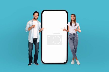 Millennial man and woman stand side by side, pointing cheerfully at a giant, blank smartphone screen, implying an interactive presentation or advertisement